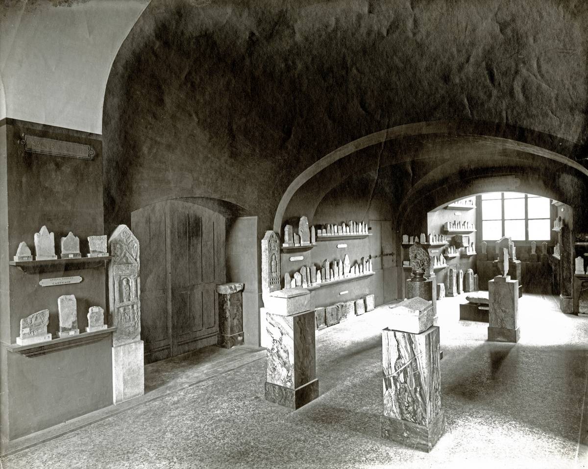 Photographie en noir et blanc montrant une ancienne salle du Louvre exposant des stèles puniques, en vue éloignée.