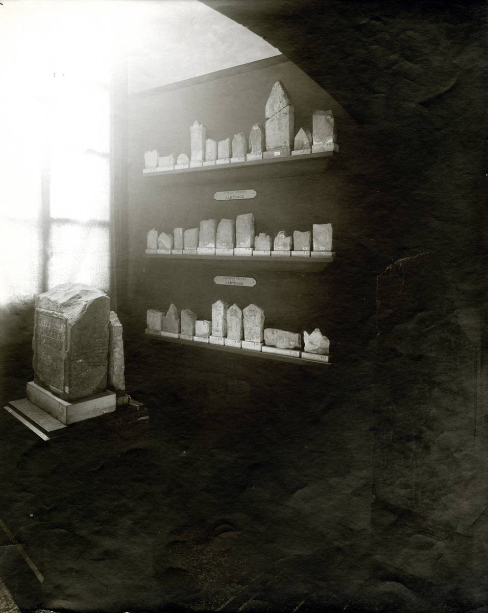 Photographie en noir et blanc montrant une ancienne salle du Louvre exposant des stèles puniques.