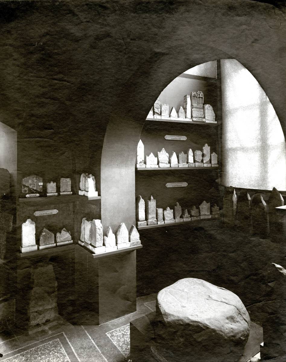 Photographie en noir et blanc montrant une ancienne salle du Louvre exposant des stèles puniques.