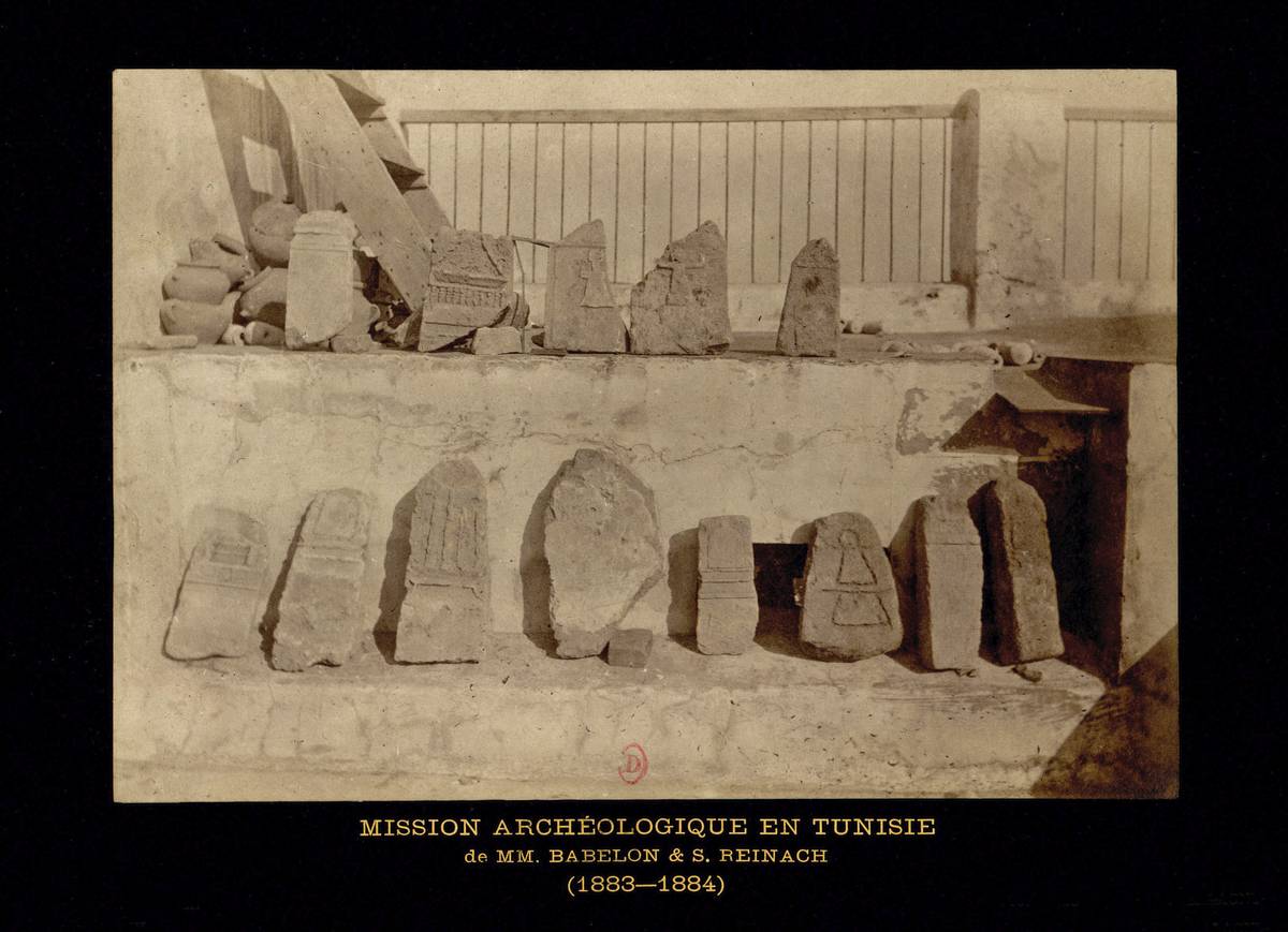 Photographie ancienne, en noir et blanc, montrant deux rangées stèles alignées le long d’un muret et posées sur lui.