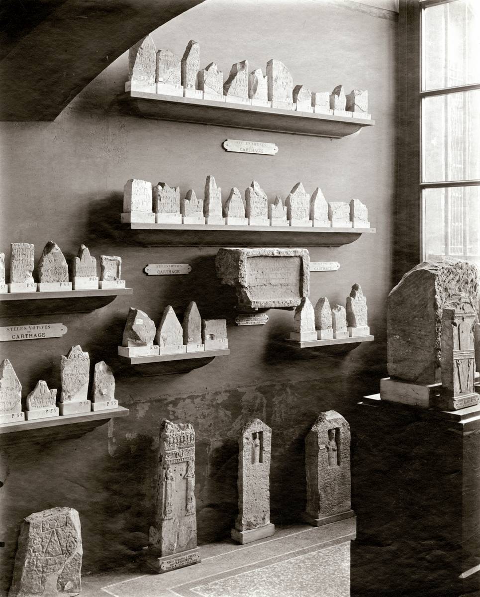 Photographie en noir et blanc montrant une ancienne salle du Louvre exposant des stèles puniques.