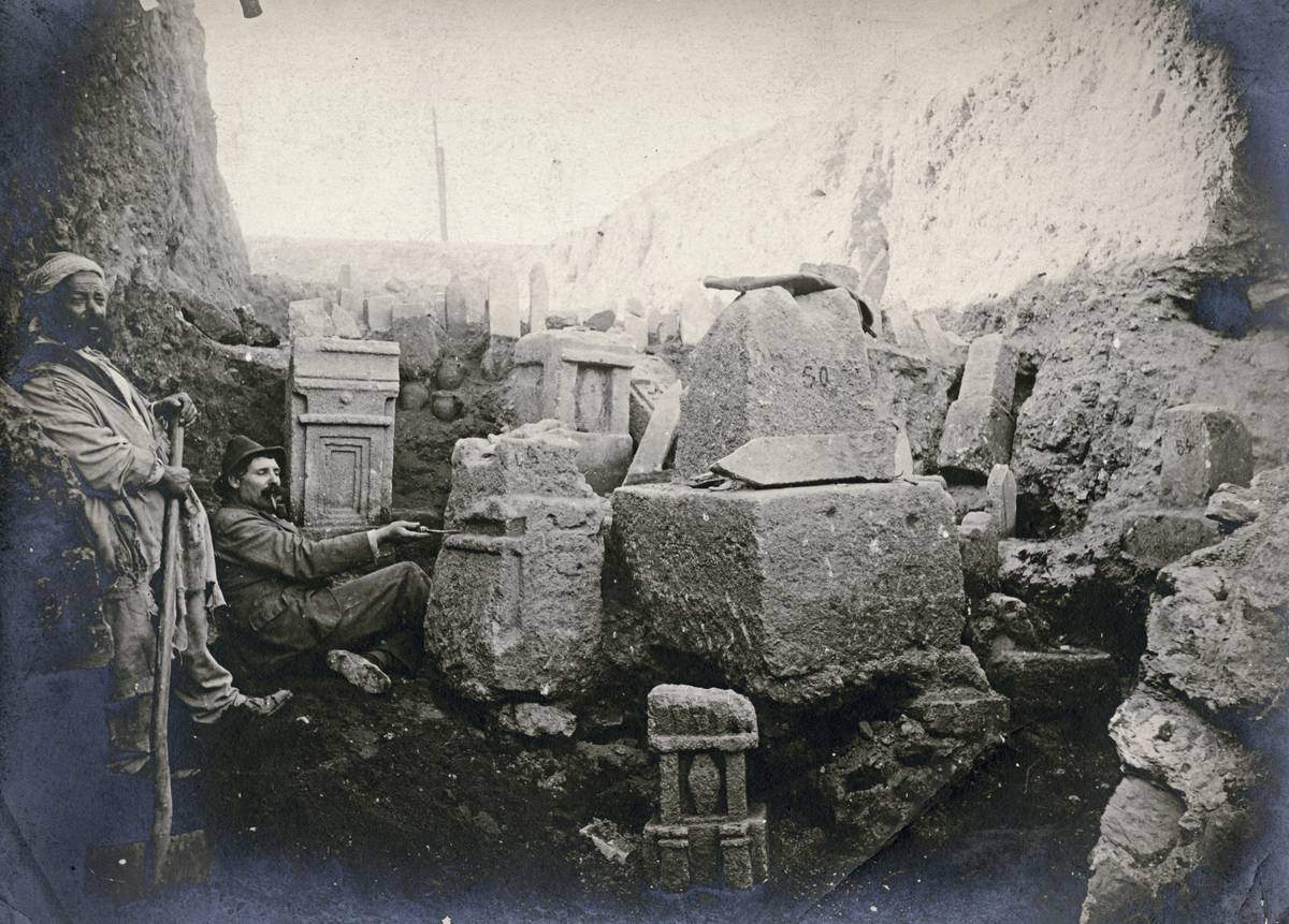 Photographie ancienne, en noir et blanc, représentant deux hommes sur un chantier de fouille, au milieu de stèles tout juste sorties de terre.