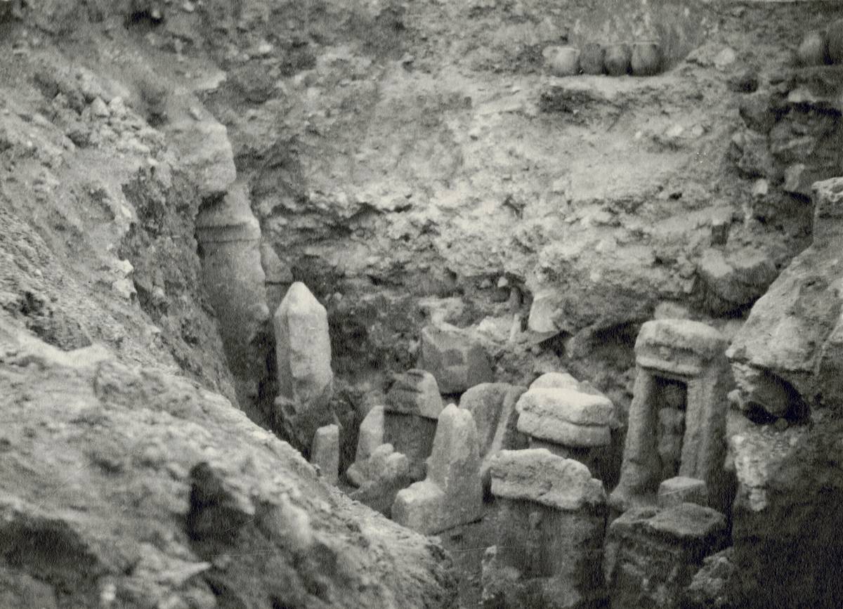 Photographie ancienne, en noir et blanc, représentant des stèles dans une zone de fouilles, entourées de monticules de terre.